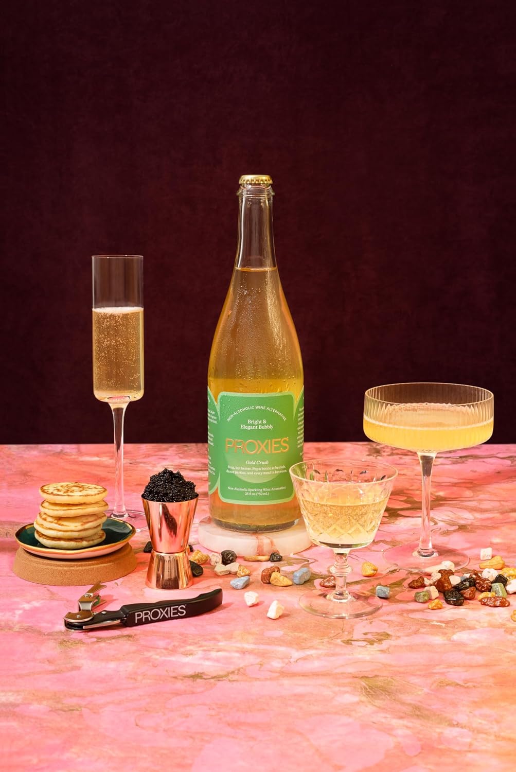 Bottle of Proxies sparkling wine with glasses and snacks on a pink surface