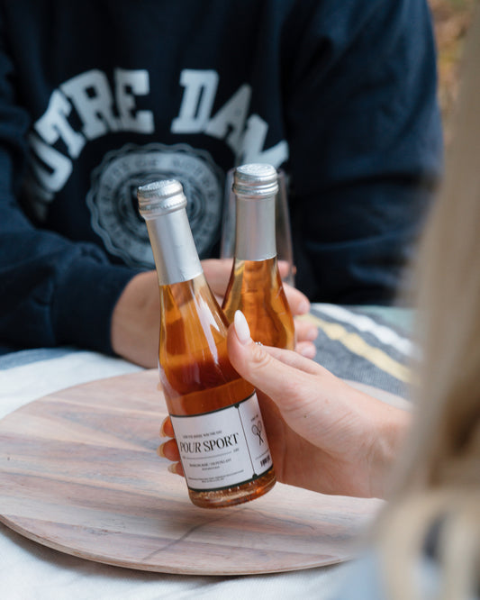 Two hands holding miniature bottles of Pour Sport on a wooden coaster.